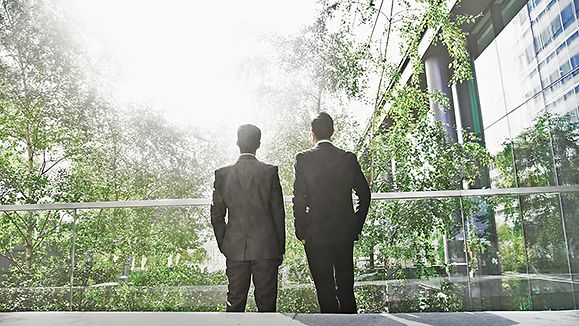 Two business men standing in green office building looking at the sun.